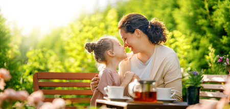 Happy little girl and her mother drinking tea in summer morning. Family sitting in the garden with cups and enjoying the conversation.の写真素材