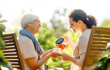 Happy young woman and her mother drinking tea in summer morning. Family sitting in the garden with cups and enjoying the conversation.の写真素材