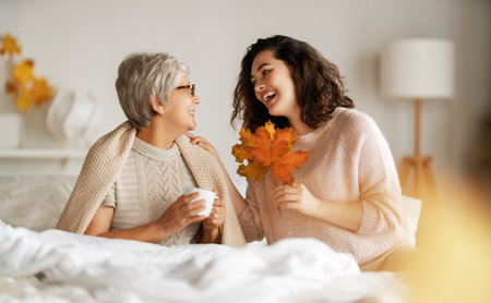 Beautiful senior mother and adult daughter are talking and smiling while sitting on bed at home.の写真素材