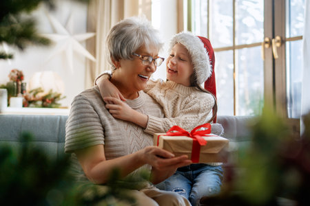 Merry Christmas and Happy Holidays! Cheerful grandma and her cute grand daughter girl exchanging gifts. Granny and little child having fun near tree indoors.の写真素材