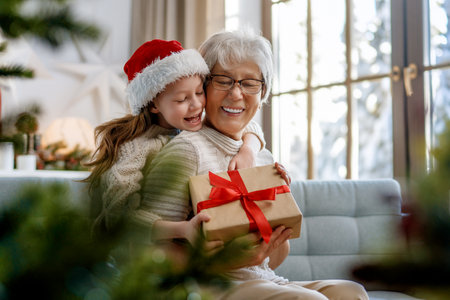 Merry Christmas and Happy Holidays! Cheerful grandma and her cute grand daughter girl exchanging gifts. Granny and little child having fun near tree indoors.の写真素材