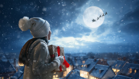 Merry Christmas. Cute little child with xmas present. Santa Claus flying in his sleigh against moon sky. Happy kid enjoy the holiday. Portrait of girl with gifts on dark background.の写真素材