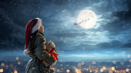 Merry Christmas. Cute little child with xmas present. Santa Claus flying in his sleigh against moon sky. Happy kid enjoy the holiday. Portrait of girl with gifts on dark background.の写真素材
