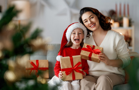 Merry Christmas and Happy Holidays. Cheerful mom and her cute daughter girl exchanging gifts. Parent and little child having fun near tree indoors.の写真素材