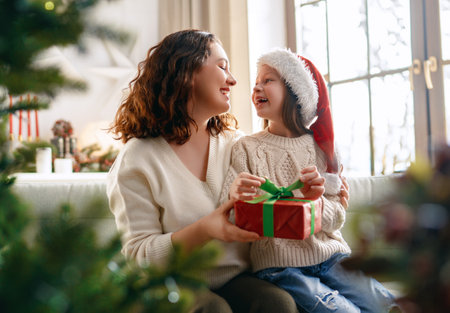 Merry Christmas and Happy Holidays. Cheerful mom and her cute daughter girl exchanging gifts. Parent and little child having fun near tree indoors.の写真素材