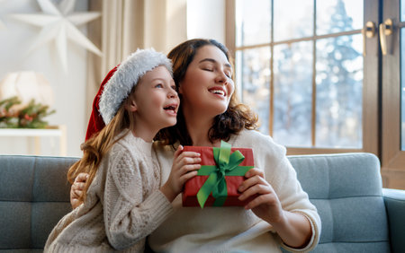 Merry Christmas and Happy Holidays. Cheerful mom and her cute daughter girl exchanging gifts. Parent and little child having fun near tree indoors.の写真素材