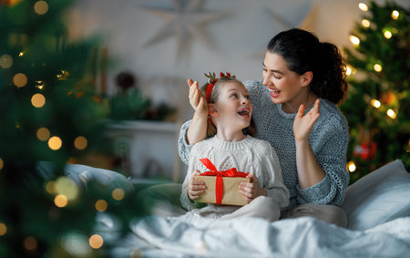 Merry Christmas and Happy Holidays. Cheerful mom and her cute daughter girl exchanging gifts. Parent and little child having fun near tree indoors. Loving family with presents in room.の写真素材