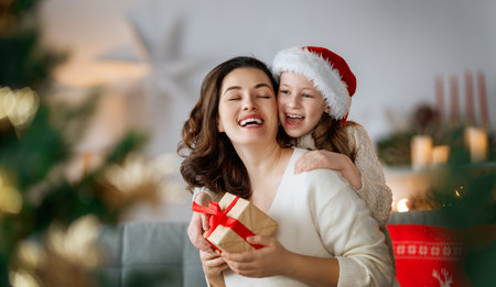 Merry Christmas and Happy Holidays. Cheerful mom and her cute daughter girl exchanging gifts. Parent and little child having fun near tree indoors.の写真素材