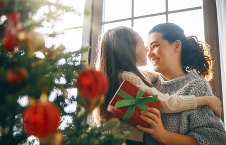 Merry Christmas and Happy Holidays. Cheerful mom and her cute daughter girl exchanging gifts. Parent and little child having fun near tree indoors.の写真素材