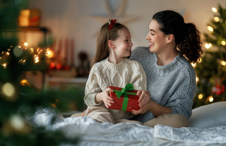 Merry Christmas and Happy Holidays. Cheerful mom and her cute daughter girl exchanging gifts. Parent and little child having fun near tree indoors. Loving family with presents in room.の写真素材