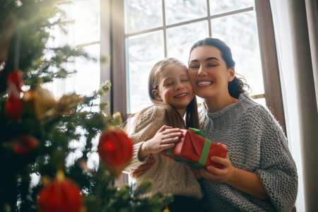 Merry Christmas and Happy Holidays. Cheerful mom and her cute daughter girl exchanging gifts. Parent and little child having fun near tree indoors.の写真素材