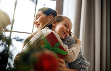 Merry Christmas and Happy Holidays. Cheerful mom and her cute daughter girl exchanging gifts. Parent and little child having fun near tree indoors.の写真素材
