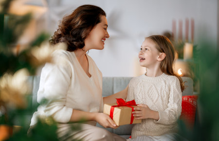 Merry Christmas and Happy Holidays. Cheerful mom and her cute daughter girl exchanging gifts. Parent and little child having fun near tree indoors.の写真素材