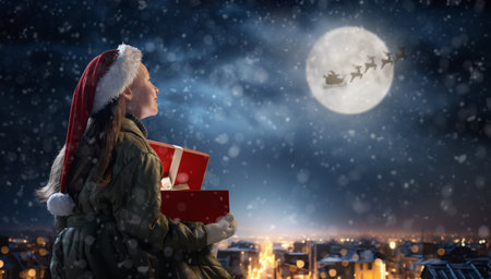 Merry Christmas. Cute little child with xmas present. Santa Claus flying in his sleigh against moon sky. Happy kid enjoy the holiday. Portrait of girl with gifts on dark background.の写真素材