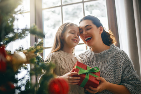 Merry Christmas and Happy Holidays. Cheerful mom and her cute daughter girl exchanging gifts. Parent and little child having fun near tree indoors.の写真素材