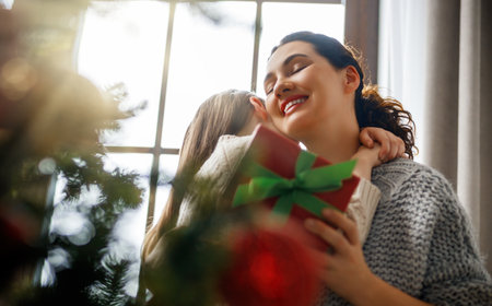 Merry Christmas and Happy Holidays. Cheerful mom and her cute daughter girl exchanging gifts. Parent and little child having fun near tree indoors.の写真素材