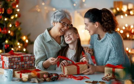 Happy Holidays. Cheerful grandmother, mother and cute daughter girl preparing for Christmas. People wrapping gifts, decorating home. Loving family with presents in room.の写真素材
