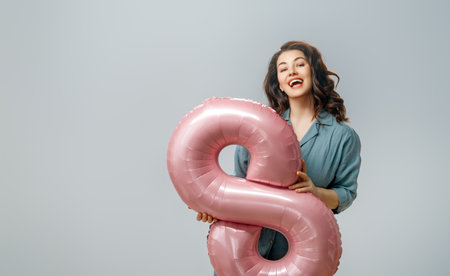 Beautiful young woman with a balloon in the shape of an eight on gray wall background.