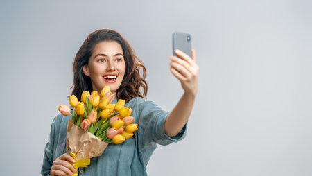 Beautiful young woman with yellow flowers in hands on grey wall background taking selfie.の写真素材