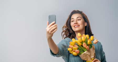 Beautiful young woman with yellow flowers in hands on grey wall background taking selfie.の写真素材