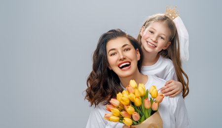 Happy women's day. Child is congratulating mom and giving her yellow flowers. Mum and girl smiling on light grey background. Family holiday and togetherness.の写真素材