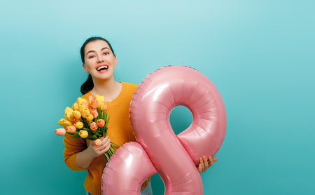 Beautiful young woman with yellow flowers and a balloon in the shape of an eight on teal wall background.の写真素材