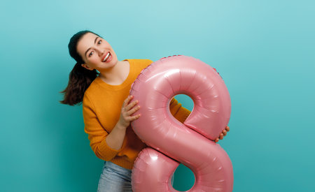 Beautiful young woman with a balloon in the shape of an eight on teal wall background.の写真素材