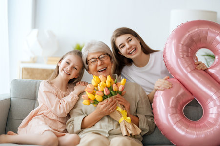 Happy mother's day! Children daughters are congratulating grandmother and giving her flowers. Granny and girls smiling and hugging. Family holiday and togetherness.の写真素材