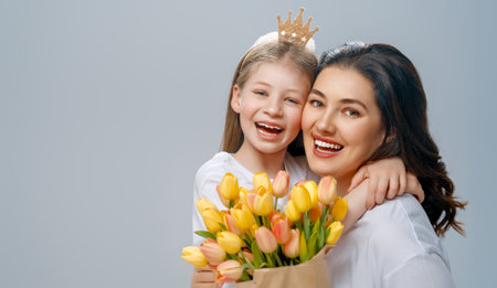 Happy women's day. Child is congratulating mom and giving her yellow flowers. Mum and girl smiling on light grey background. Family holiday and togetherness.の写真素材