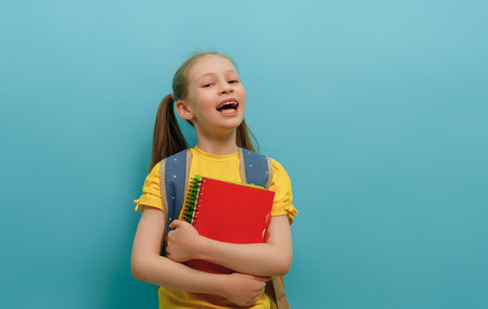 Cute happy kid standing on light blue background. Child with backpack, little girl is ready to back to school.の写真素材