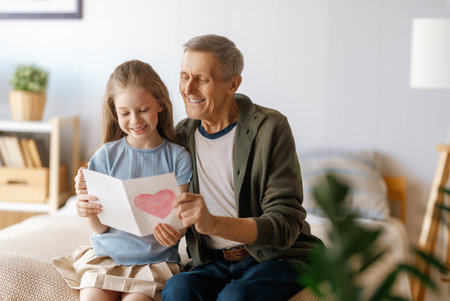 Child granddaughter is congratulating her grandfather and giving him postcard. Girl is smiling. Family holiday and togetherness.の写真素材