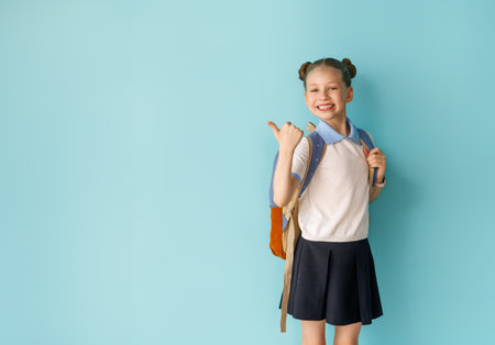 Cute happy teenager standing on bright light blue background. Child with backpack, girl is ready to...