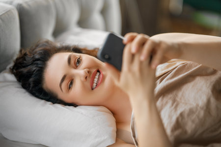 Happy casual beautiful woman is using a phone in the bed at home.の写真素材