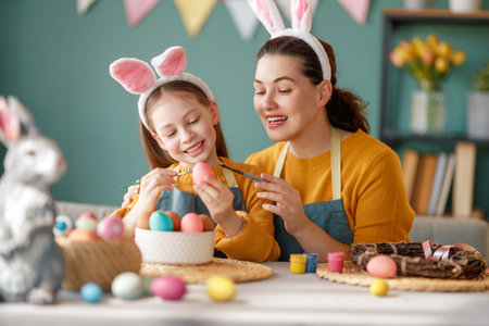 Mother and her daughter painting eggs. Happy family preparing for Easter. Cute little child girl wearing bunny ears.の写真素材