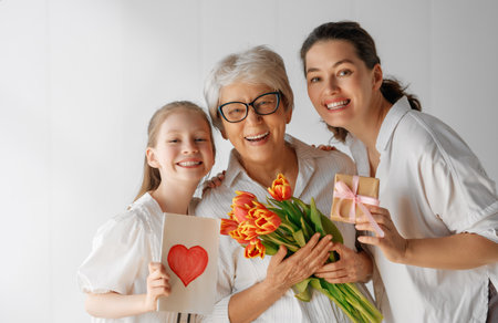 Happy mother's day! Child daughter is congratulating mom and grandma giving them flowers tulips. Granny, mom and girl smiling on light grey background. Family holiday and togetherness.の写真素材