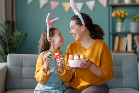 Happy holiday! Mother and her daughter with painting eggs. Family celebrating Easter. Cute little child girl is wearing bunny ears.の写真素材