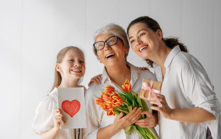 Happy mother's day! Child daughter is congratulating mom and grandma giving them flowers tulips. Granny, mom and girl smiling on light grey background. Family holiday and togetherness.の写真素材