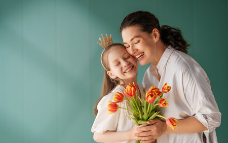 Child daughter is congratulating mom and giving her flowers tulips. Mom and girl smiling on green background. Family holiday and togetherness.の写真素材