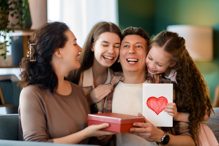 Children daughters are congratulating father and giving him postcard and gift. Dad, mom and girls are smiling and hugging. Family holiday and togetherness.の写真素材