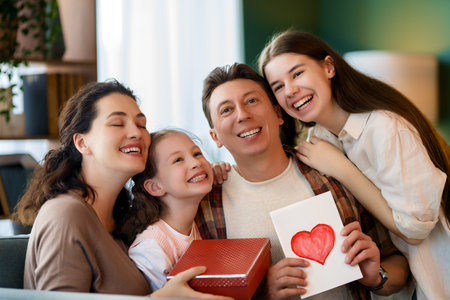 Children daughters are congratulating father and giving him gift. Dad, mom and girls are smiling and hugging. Family holiday and togetherness.の写真素材