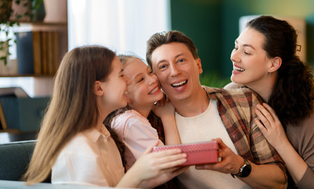 Children daughters are congratulating father and giving him gift. Dad, mom and girls are smiling and hugging. Family holiday and togetherness.の写真素材