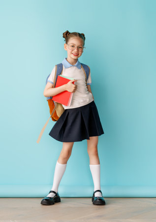 Cute happy teenager standing on bright light blue background. Child with backpack, girl is ready to back to school.の写真素材