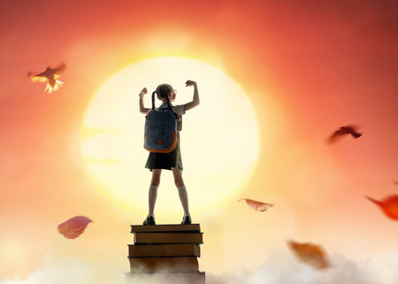 Back to school. Happy cute industrious child standing on the tower of books on background of sunset sky. Concept of education and reading. The development of the imagination.の写真素材
