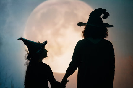 Woman and her daughter dressed in witch costumes for Halloween. Family standing on full moon background.の写真素材