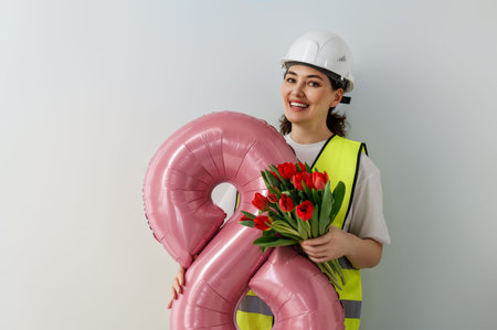 A cheerful female construction worker, wearing a helmet and safety vest, holding a pink balloon in the shape of the number eight and a bouquet of vibrant red tulips. This image embodies celebration, positivity, and empowerment in the workplace. Perfect for concepts related to Women's Day, construction themes, and workplace diversity.の写真素材