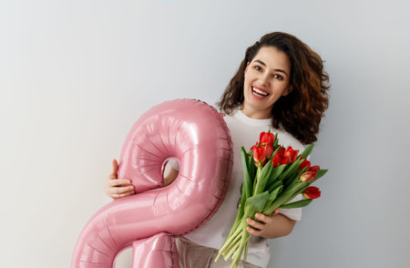 Joyful young woman smiling brightly while holding a large pink figure eight balloon and a bouquet of...