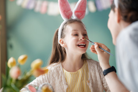 A happy young girl wearing fluffy pink bunny ears smiles warmly as her nose is painted by an adult, capturing the playful joy of Easter festivities. The soft colors, whimsical background, and fresh spring flowers evoke feelings of family bonding, creativity, and the delight of childhood traditions. Perfect for commercial, editorial, and lifestyle concepts focusing on holiday celebrations, family moments, and joyful springtime activities.の写真素材