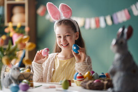 Joyful young girl wearing pink bunny ears headband smiles brightly while holding painted Easter eggs...