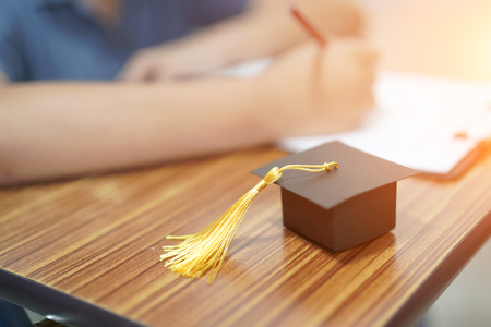 Asian children young kid write answer in a quiz examination test study learn with graduation gap hat on wooden table in school : bright genius education concept.の写真素材