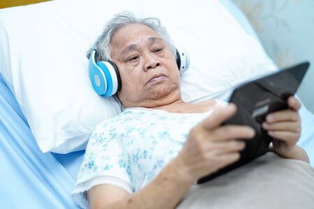Asian senior or elderly old lady woman patient use earphone while lie down and happy on bed in nursing hospital ward, healthy strong medical conceptの写真素材